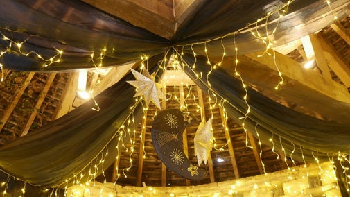 The timber frame ceiling of Avebury's 16th Century Dovecote decorated with black drapes, hanging paper star ornaments, warm fairy lights, and a crescent moon decoration featuring golden star and comet designs.